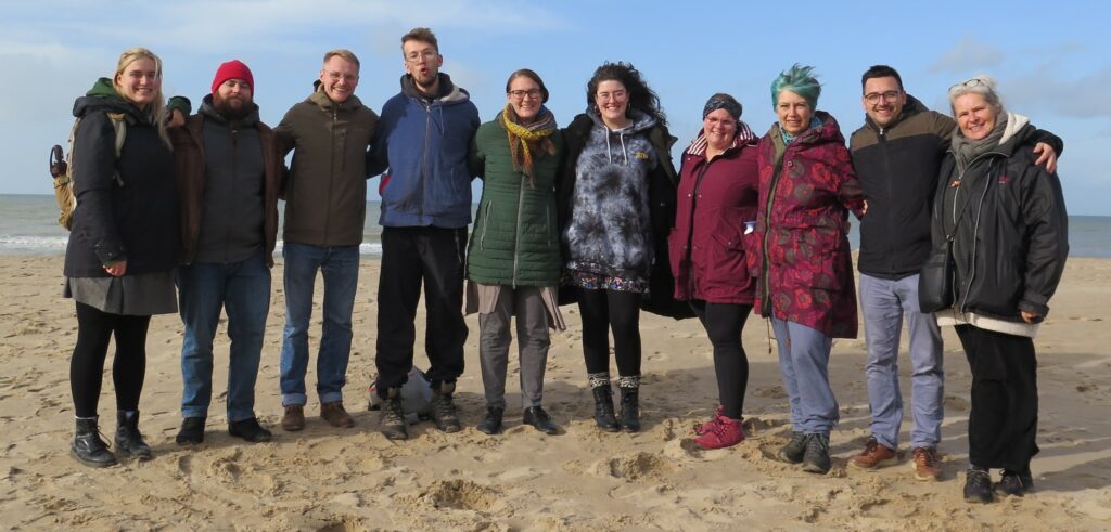 Das Vokalensemble Piece of Peace steht Arm in Arm an einem Strand. Es ist ruppig kalt, wie man an ihrer Winterbekleidung sieht.