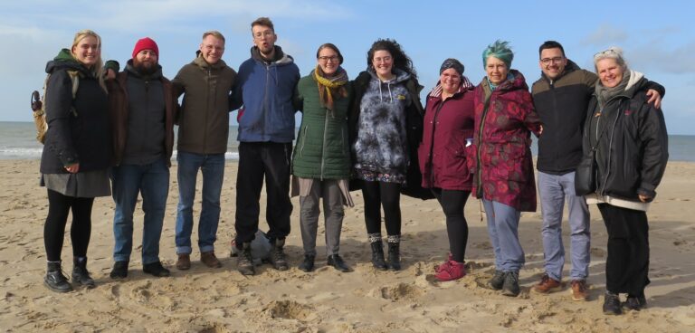 Das Vokalensemble Piece of Peace steht Arm in Arm an einem Strand. Es ist ruppig kalt, wie man an ihrer Winterbekleidung sieht.